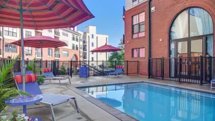 A sparkling outdoor swimming pool with surrounding lounge chairs, red umbrellas, and a view of the modern apartment buildings.