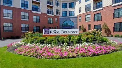 The front entrance of The Royal Belmont Luxury Apartments with blooming flower beds and a welcoming sign.