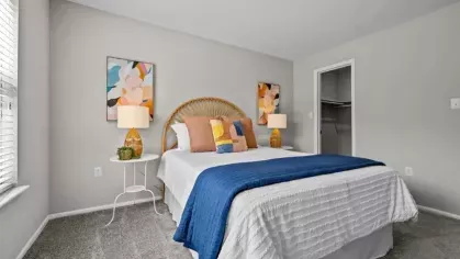 A cozy bedroom with a rattan headboard, white bedding accented with a navy blue throw, and colorful abstract artwork above matching bedside lamps.