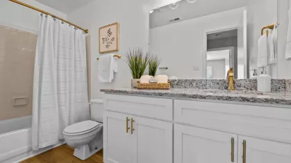 A modern bathroom featuring a granite countertop, white vanity with gold hardware, a large mirror with stylish lighting, and a bathtub with a white shower curtain.