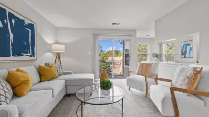 A bright living room with a gray sectional sofa, mustard yellow accent pillows, white armchairs, and a sliding glass door leading to a balcony with a scenic view.