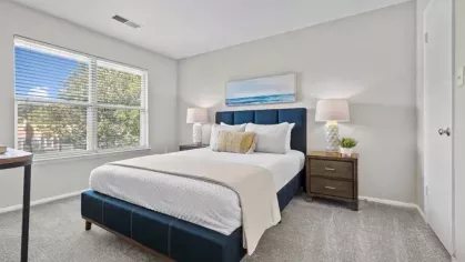 A stylish bedroom with a navy blue upholstered bed, white and beige bedding, matching bedside tables with lamps, and a large window providing natural light.