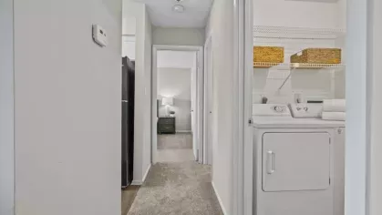 A hallway with soft carpeting leading to a bedroom, featuring a built-in laundry area with a washer, dryer, and storage shelves.