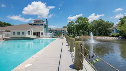 A resort-style pool area with a wooden boardwalk leading to a serene pond with a water fountain, surrounded by lush greenery.