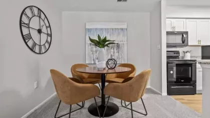 A dining nook with a round wooden table, brown upholstered chairs, a large decorative wall clock, and an adjacent modern kitchen.