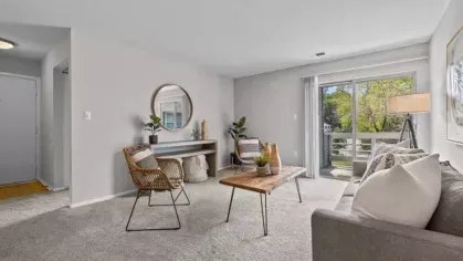 A spacious living room with a gray couch, a wooden coffee table, rattan chairs, and a large sliding door leading to a balcony with greenery.