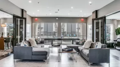 A stylish resident lounge with modern gray sofas, marble flooring, and large windows offering a view of the outdoor pool area.