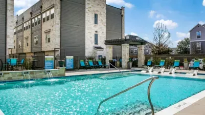 A sparkling outdoor pool with lounge chairs, surrounded by stone-accented buildings and a bright blue sky.