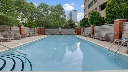 A spacious outdoor pool area with modern seating, trees, and the apartment buildings in the background.