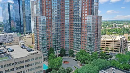 An aerial view showcasing the towering red-brick and glass Skyline New Rochelle apartments surrounded by lush trees and urban scenery.