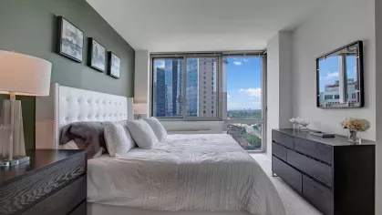 A luxurious bedroom featuring a tufted white headboard, large windows with urban views, and sophisticated decor, providing a tranquil retreat.