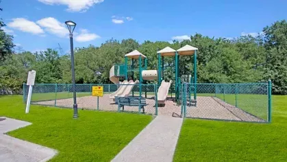 Fenced playground with green and beige play structures surrounded by lush greenery.