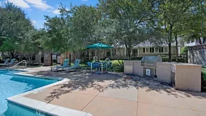Outdoor poolside seating area with barbecue grills and shaded tables under green umbrellas.