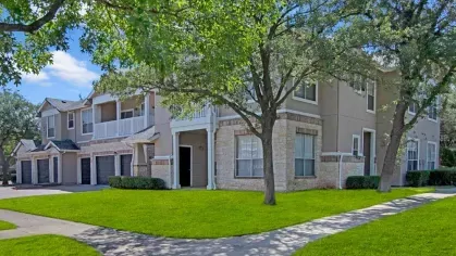 Two-story beige apartment building with stone accents, surrounded by lush green lawns and trees.
