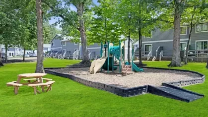 Shaded playground with slides and climbing equipment surrounded by mature trees and green lawns.