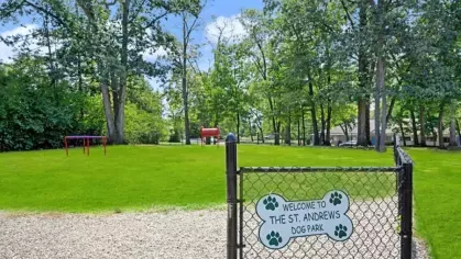 Fenced dog park with a welcoming sign, lush greenery, and open space for pets to play.