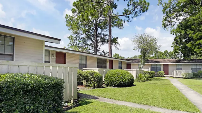 Row of beige single-story homes with red doors, white fencing, and neatly trimmed bushes.