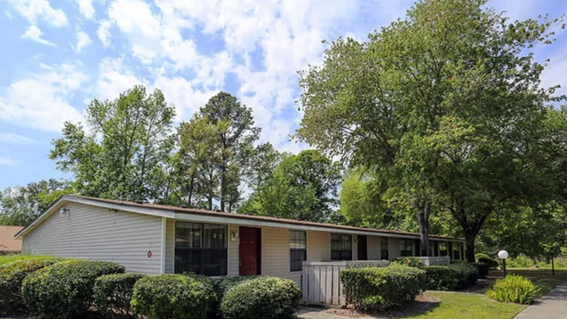 Beige house with a red door and surrounding greenery, set against a backdrop of tall trees and blue sky.