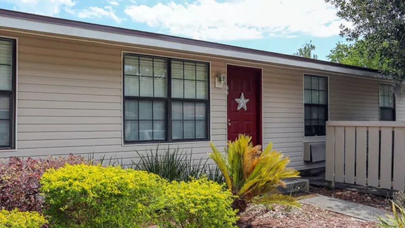 Beige house with a bold red door, surrounded by vibrant green and yellow landscaping.