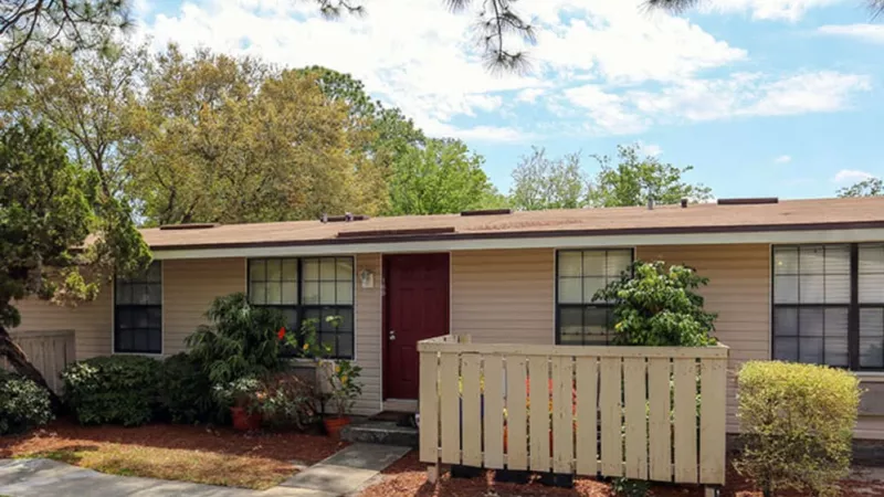 Beige home with a red door, white picket-style fencing, and lush landscaping.
