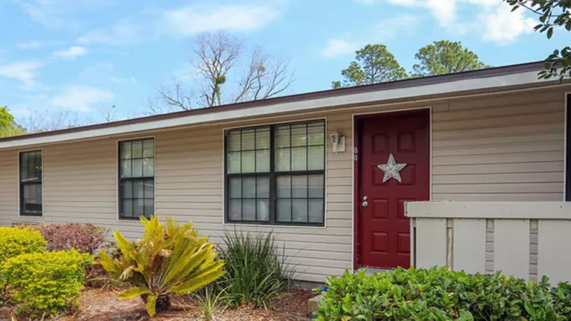 Beige house with a red door adorned with a silver star, surrounded by well-maintained plants and shrubs.