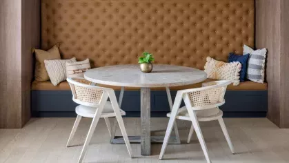 A cozy dining nook at Vista At Town Green, featuring a round wooden table with two woven chairs, a tufted leather bench, and decorative pillows set against a floral-patterned accent wall.