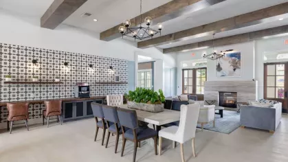A stylish and spacious clubhouse at Vista At Town Green, offering a mix of modern and rustic design with exposed beams, a statement chandelier, a coffee bar, and comfortable lounge seating near a fireplace.