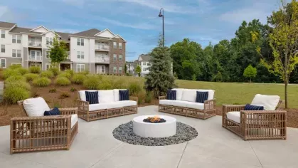 A beautifully designed outdoor fire pit lounge with wicker seating and plush cushions, offering a serene gathering space with lush landscaping and apartment buildings in the background.