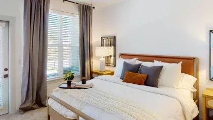 Spacious bedroom with a modern design, featuring a plush bed with layered pillows, bedside tables, and soft natural light through large windows.