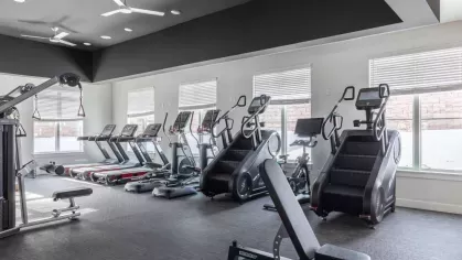Spacious gym area featuring treadmills, stair climbers, and cardio equipment with large windows offering natural light.