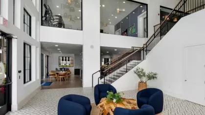 A modern lobby with high ceilings, blue accent chairs, and a staircase leading to an open mezzanine.