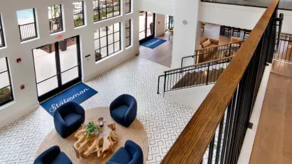 An airy lobby view from above, showcasing blue chairs on a circular rug and large windows with natural light.