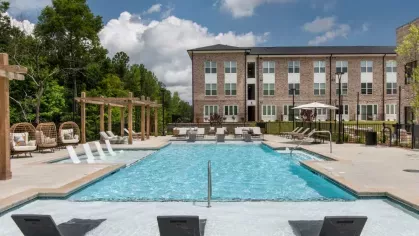 A sparkling pool area with modern loungers, pergola seating, and a backdrop of brick apartment buildings.