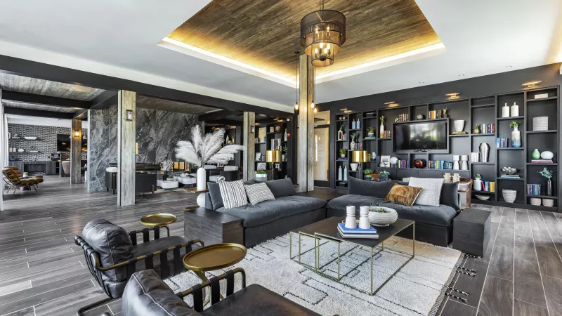 The stylish entertainment lounge at The Warner Apartments features a curved mustard-colored sofa, natural wood coffee tables, and a sleek bar area with industrial pendant lighting.