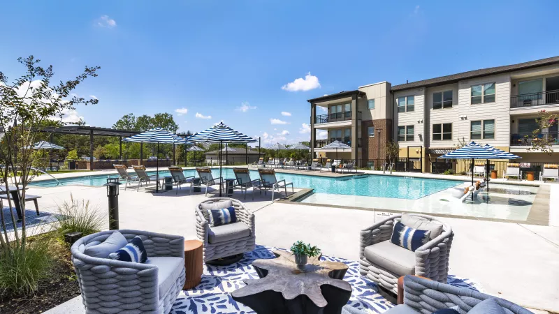 The luxury pool area features stylish woven chairs, a sculptural table, and blue-patterned umbrellas, offering a perfect spot for relaxation.