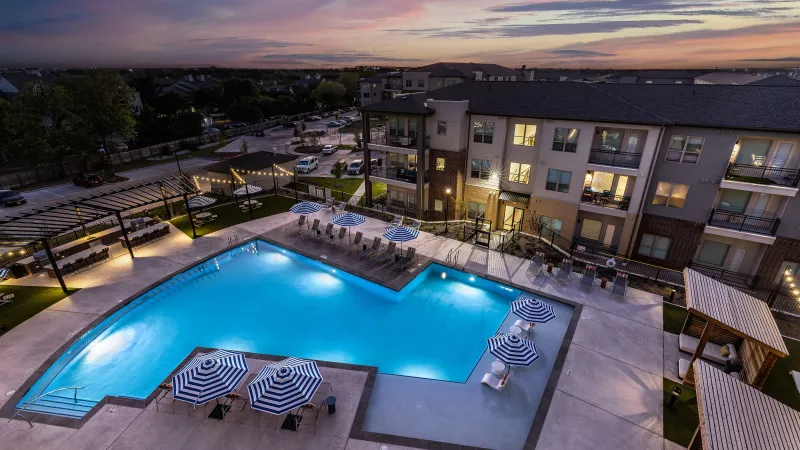 The illuminated pool glows under the evening sky, surrounded by stylish seating, pergolas, and lush landscaping.