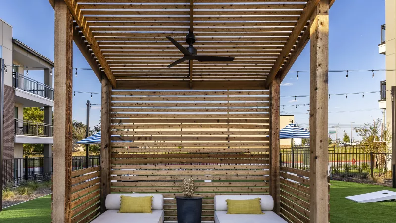 A wooden pergola shelters two plush daybeds with accent pillows, offering a peaceful retreat by the pool.