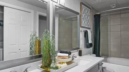 A modern bathroom featuring a long vanity with dual sinks, a large mirror, and a clean white bathtub with gray tile accents.