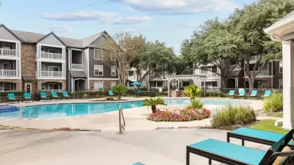 A sparkling resort-style pool with bright blue lounge chairs and surrounding apartment buildings under a sunny sky.