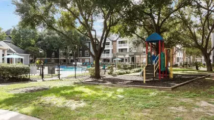 A shaded playground with colorful equipment, green grass, and a view of the pool area and apartments.
