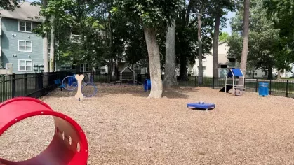 A fenced dog park with agility equipment, trees for shade, and woodchip ground cover, surrounded by apartment buildings.