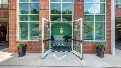 A welcoming entrance featuring a modern brick facade, glass double doors, and elegant planters, leading to a stylish lobby with a grand piano and bold geometric flooring.