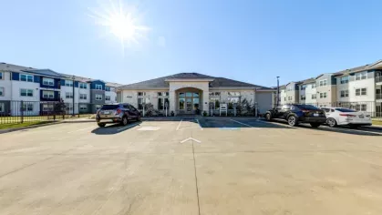 A bright exterior view of the Linden Ranch Luxury Apartments clubhouse with a stone facade, large arched entrance, and adjacent modern apartment buildings under a clear blue sky.