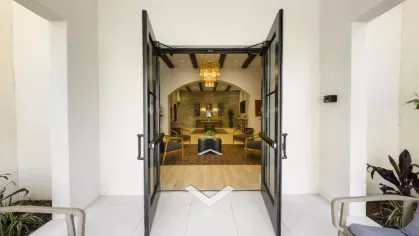 A welcoming entrance to Locklyn West Palm Luxury Apartments featuring stylish black-framed glass doors opening into a beautifully designed lobby with warm lighting, a modern chandelier, and comfortable seating.