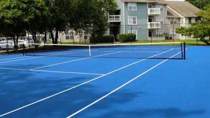 A vibrant blue tennis court with white lines and a black net, surrounded by apartment buildings and lush trees.