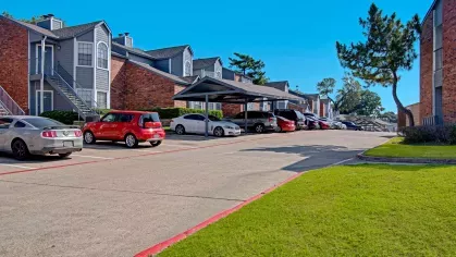 Parking lot with covered and uncovered spaces in front of two-story red brick and gray siding apartment buildings, with neatly trimmed lawns and a clear blue sky.