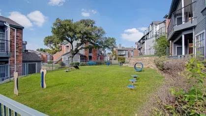 Fenced-in dog park with agility equipment and grassy play area between apartment buildings