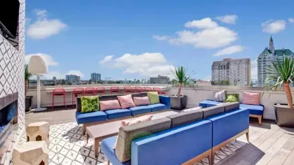 A rooftop deck with colorful seating, lush plants, and a mix of tables and lounge chairs overlooking the city.