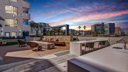 A rooftop lounge at sunset with modern seating, an outdoor kitchen, and a fire pit, overlooking the city skyline.