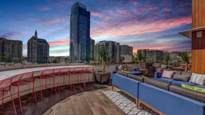 A rooftop lounge with plush blue sofas, red bar stools, and panoramic city views under a vibrant sunset sky.